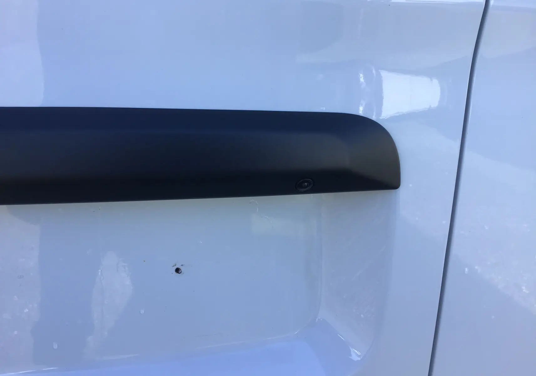 Gros plan sur la caméra de recul intégrée dans la protection latérale noire du côté gauche du FIAT Scudo Fourgon blanc.