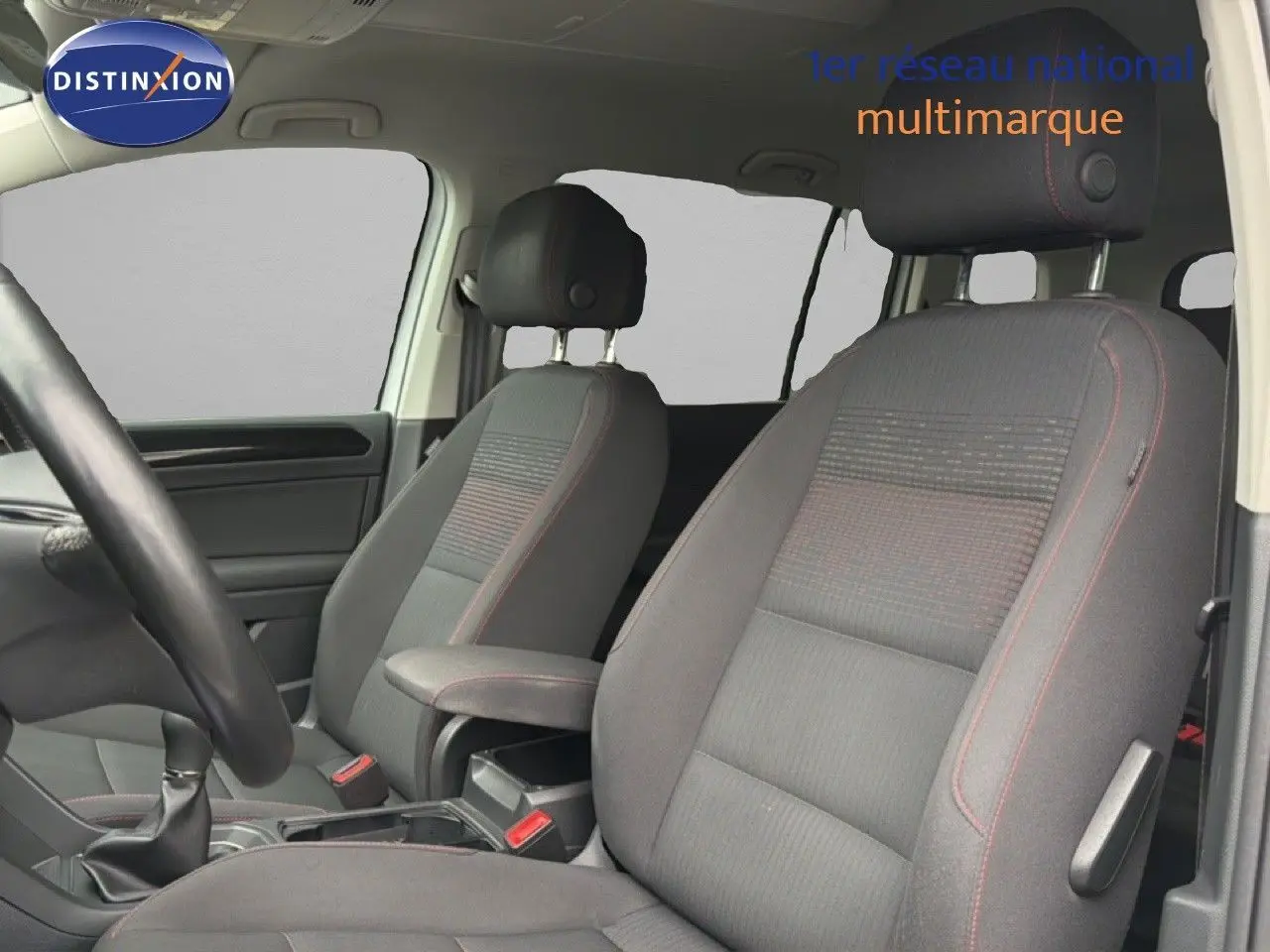 Vue intérieure côté conducteur du Volkswagen Touran blanc de 2018, montrant les sièges avant en tissu gris avec surpiqûres rouges.