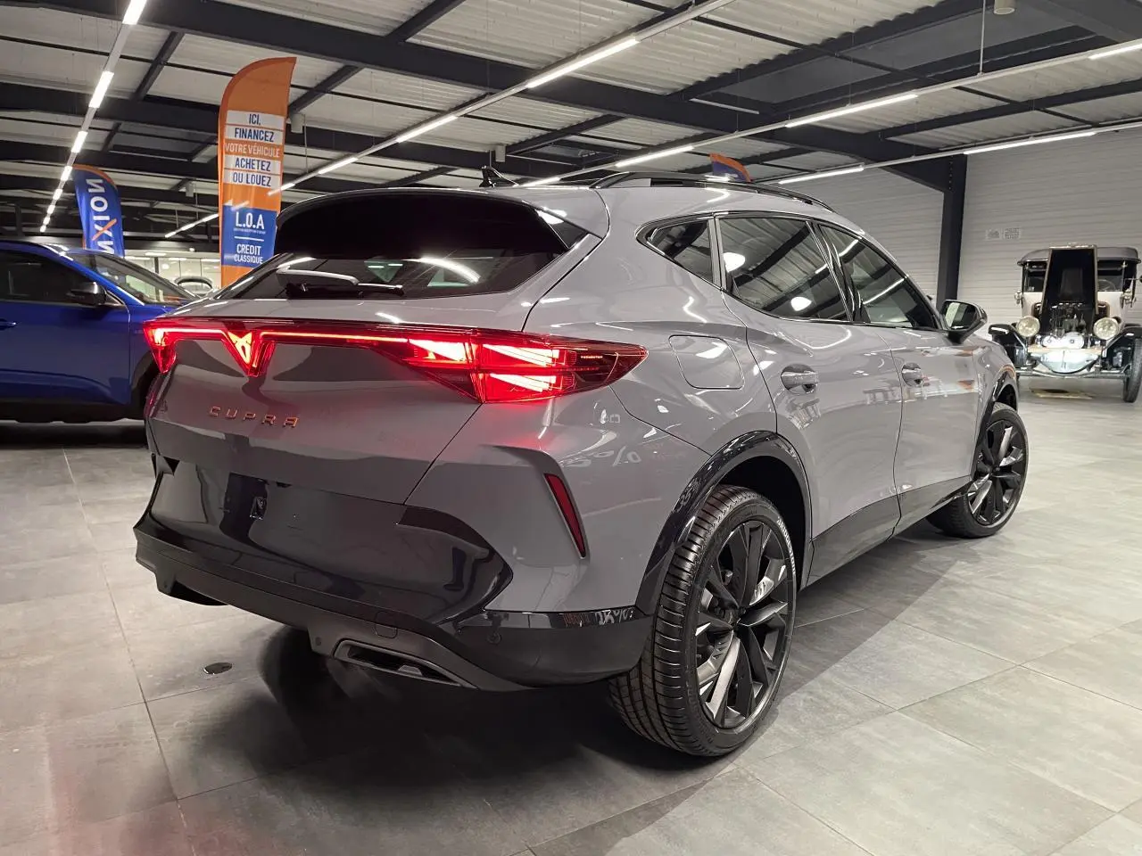 Vue 3/4 arrière droite du CUPRA Formentor gris Graphene avec feux arrière allumés et jantes alliage noires en showroom.