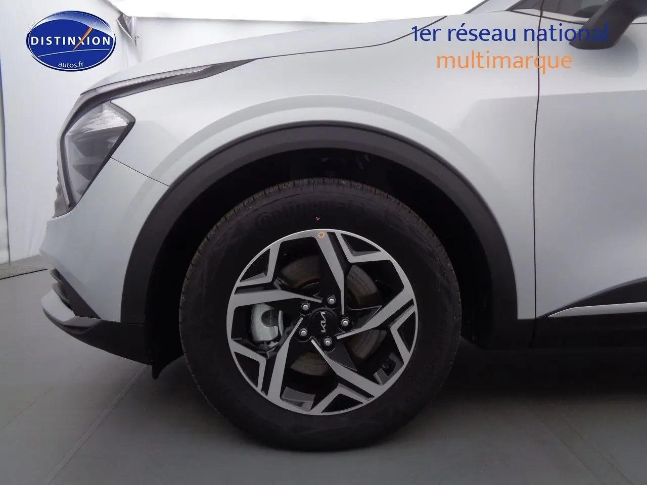 Gros plan sur la roue avant gauche et l'aile d'un KIA Sportage gris acier métal, avec jante bicolore et phares partiellement visibles.