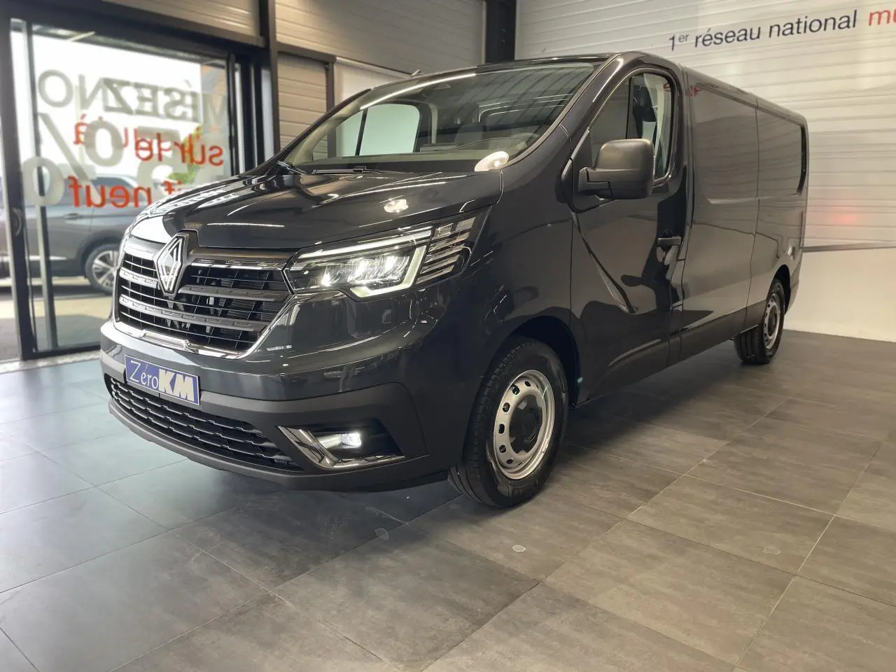 Vue 3/4 avant droit d'un Renault Trafic Fourgon gris comète avec phares LED et jantes tôles 16 pouces en showroom.