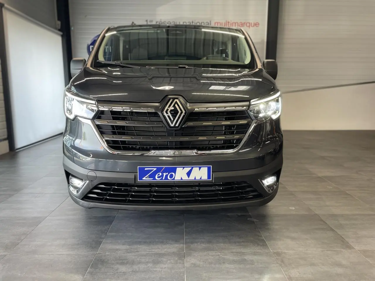 Vue frontale d'un Renault Trafic Fourgon gris Comète avec phares et feux de jour allumés en intérieur showroom.
