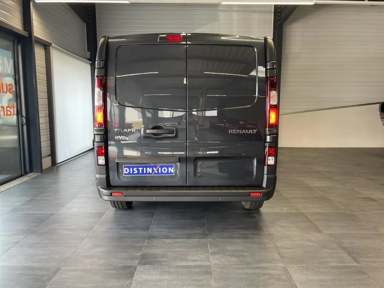 Vue arrière du Renault Trafic Fourgon gris Comète avec feux allumés et plaque Distinxion visible en intérieur showroom.