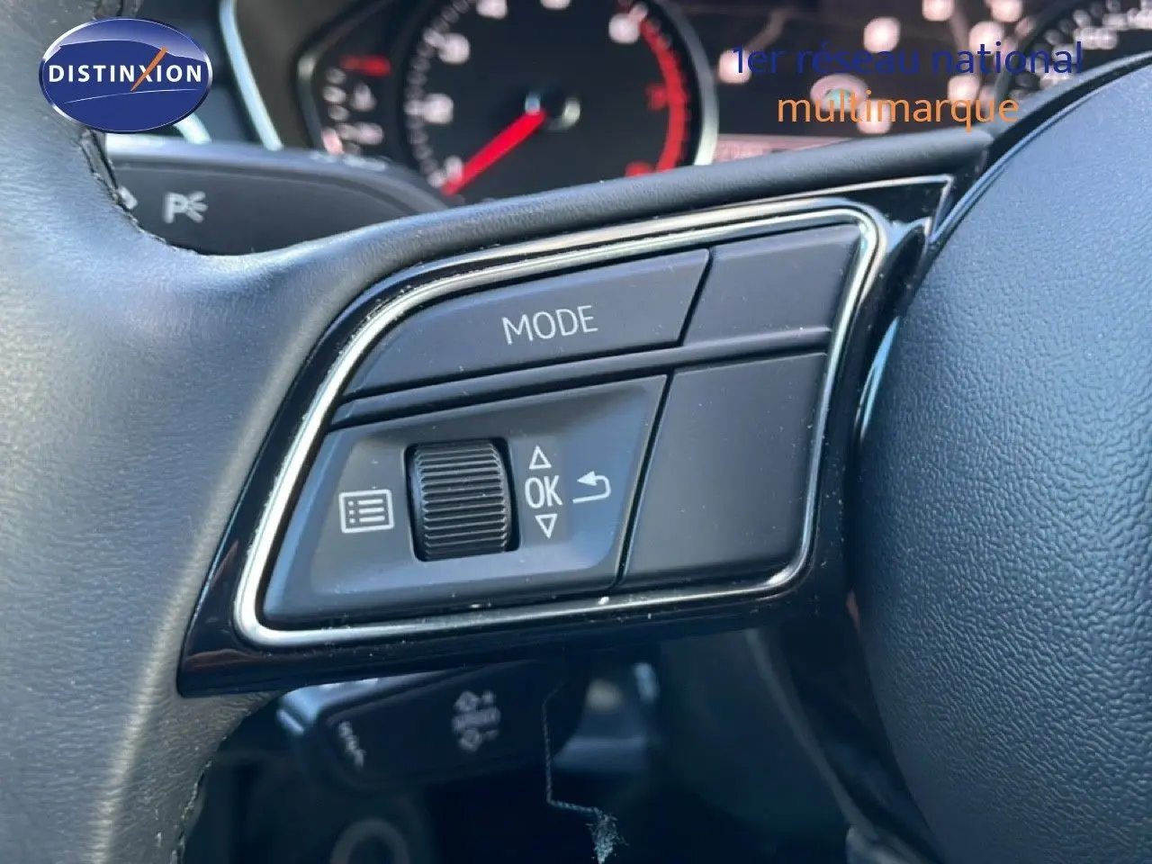 Gros plan sur les commandes gauche du volant noir de l'Audi A4 Avant gris Manhattan, avec tableau de bord flou en arrière-plan.