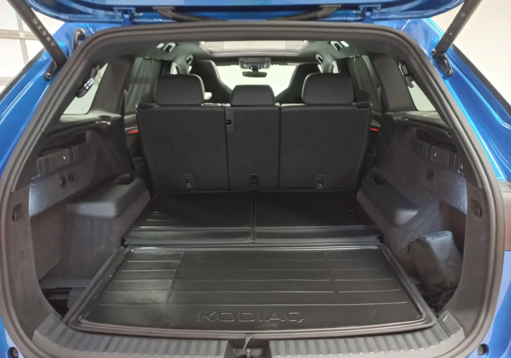Coffre ouvert vu de l'arrière d'un Skoda Kodiaq Bleu RS 2024 avec banquette arrière noire et tapis de coffre marqué KODIAQ.