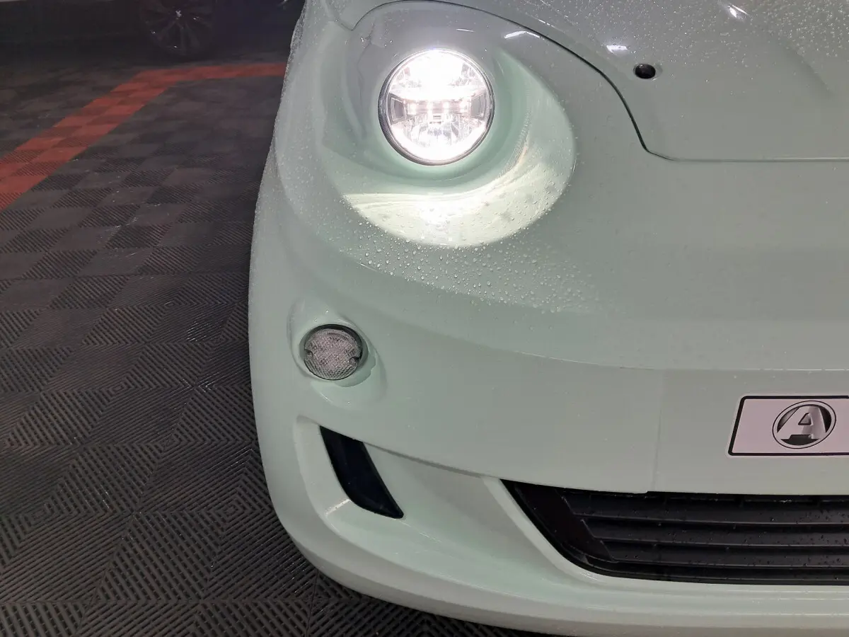 Gros plan sur l'avant gauche de l'AIXAM MINAUTO E chic 2026 vert électrique avec phare allumé et gouttes d'eau visibles.