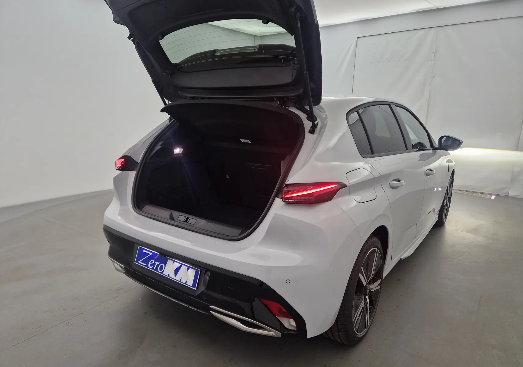 Vue 3/4 arrière droite d'une Peugeot 308 blanche Okenite avec coffre ouvert et feux arrière allumés dans un studio.