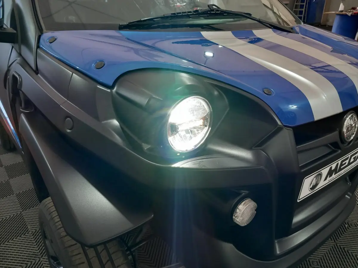 Gros plan sur l’avant gauche bleu électrique de l’AIXAM MEGA E-SCOUTY EVO avec phare allumé et bandes blanches sur le capot.