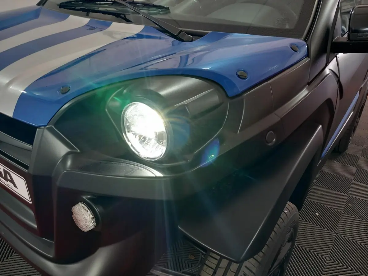 Gros plan avant droit sur l'AIXAM MEGA bleu électrique avec phare allumé et rétroviseur caméra côté conducteur.