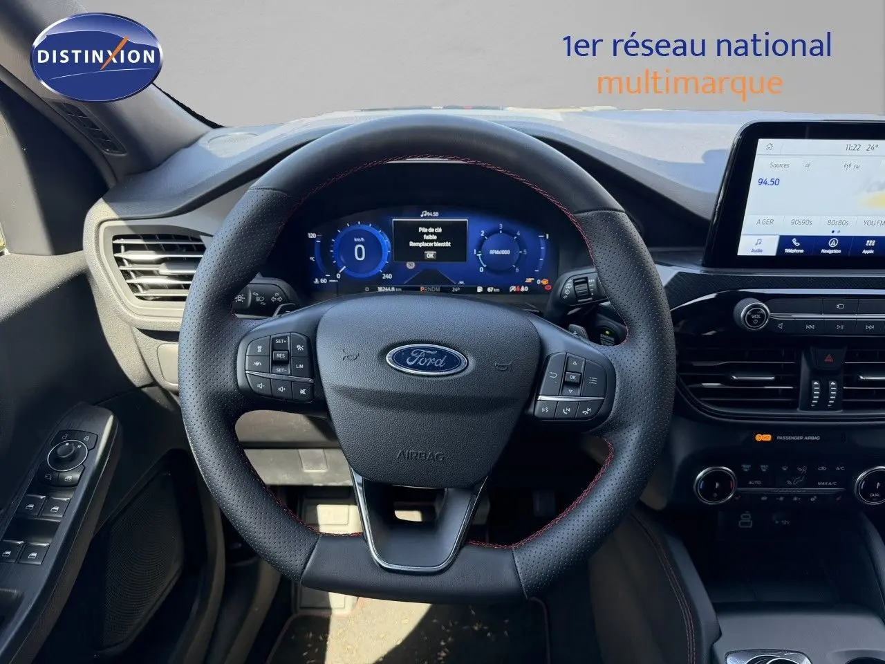 Vue intérieure centrée sur le volant noir perforé avec surpiqûres rouges du Ford Kuga 2024, tableau de bord digital et écran tactile.