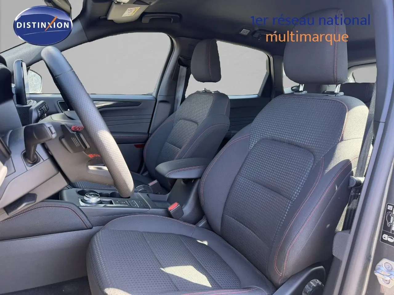 Vue intérieure côté conducteur du Ford Kuga 2024, sièges tissu noir avec surpiqûres rouges et console centrale moderne.