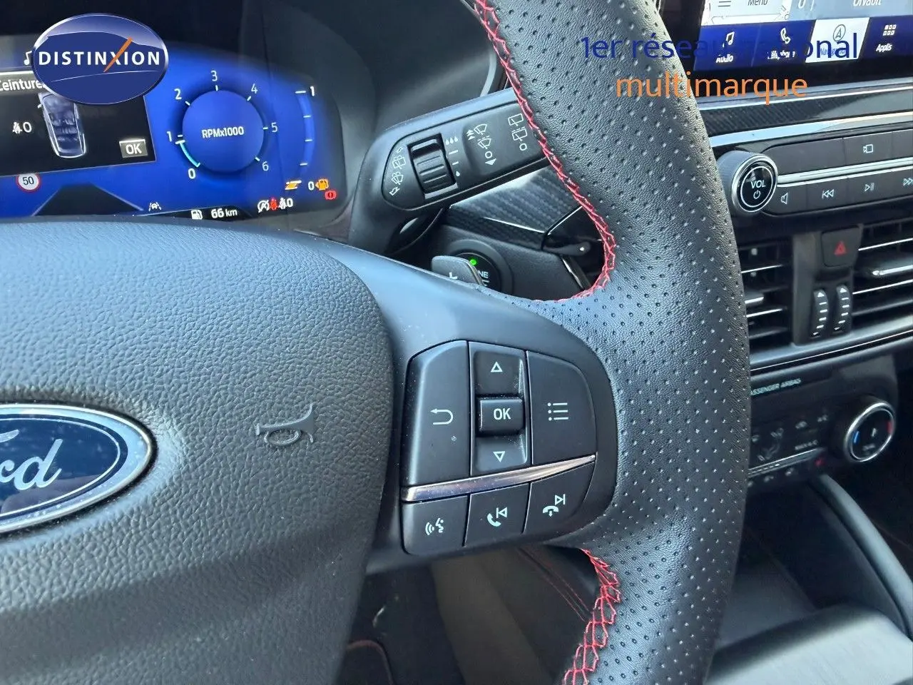 Gros plan sur le volant en cuir perforé avec surpiqûres rouges du Ford Kuga 2024, tableau de bord digital bleu en arrière-plan.