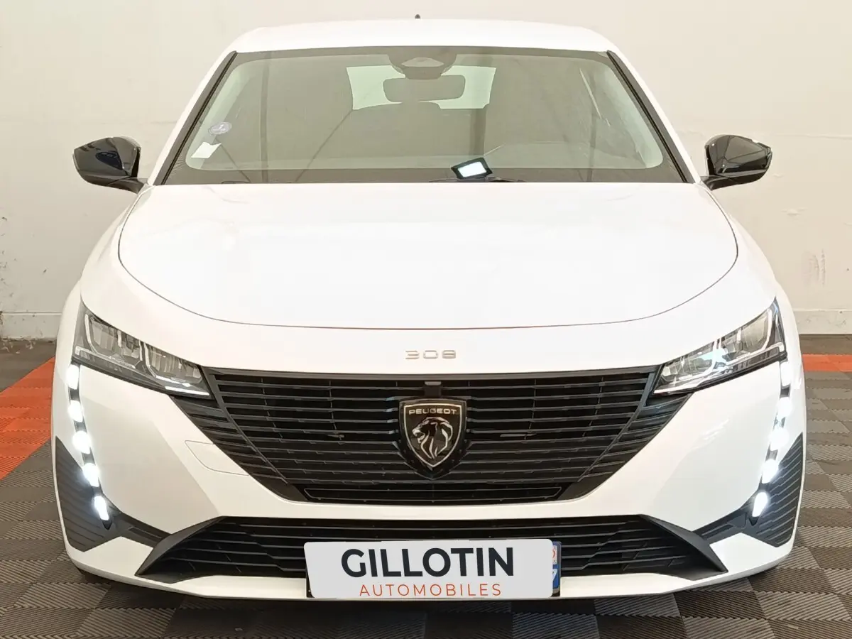 Vue frontale d'une Peugeot 308 blanche 2023 avec calandre noire et feux LED verticaux allumés.