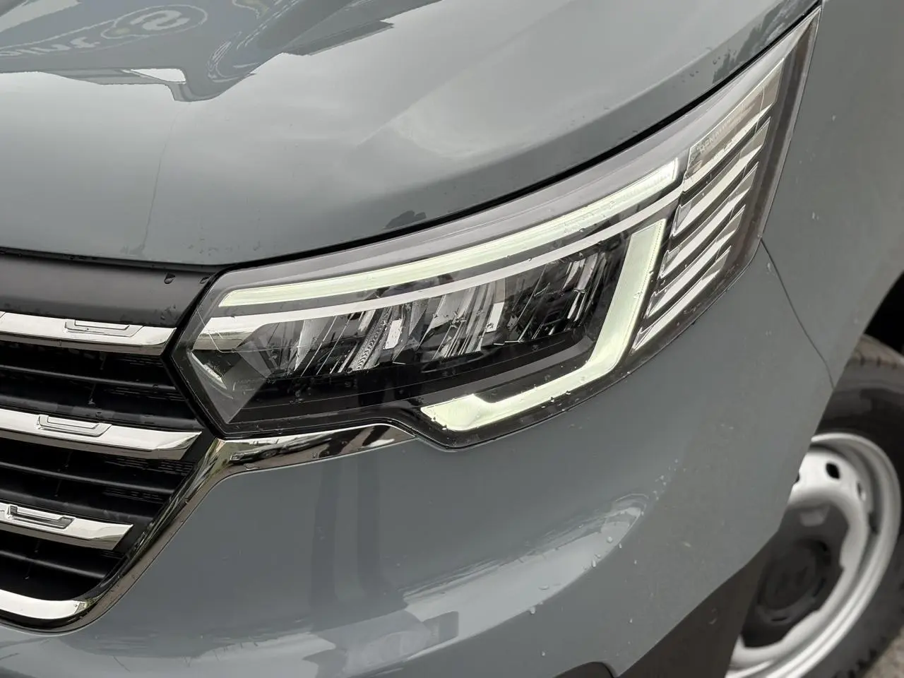 Gros plan sur le phare avant droit du Renault Trafic Fourgon gris urbain, soulignant la calandre chromée et la roue acier.