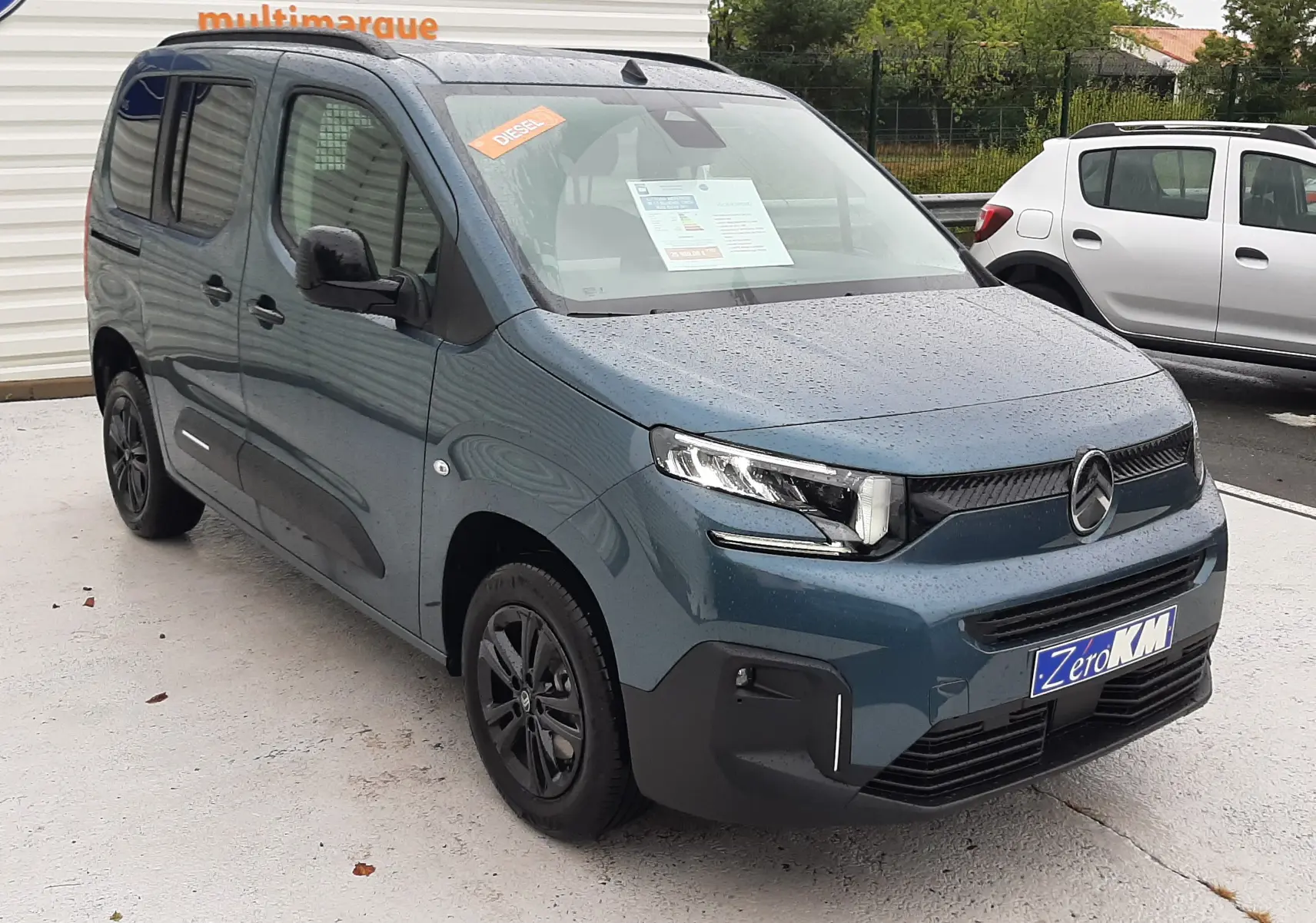 Vue 3/4 avant gauche d'un Citroën Berlingo bleu Kiama avec stickers Diesel et plaque Zero KM sous la pluie.