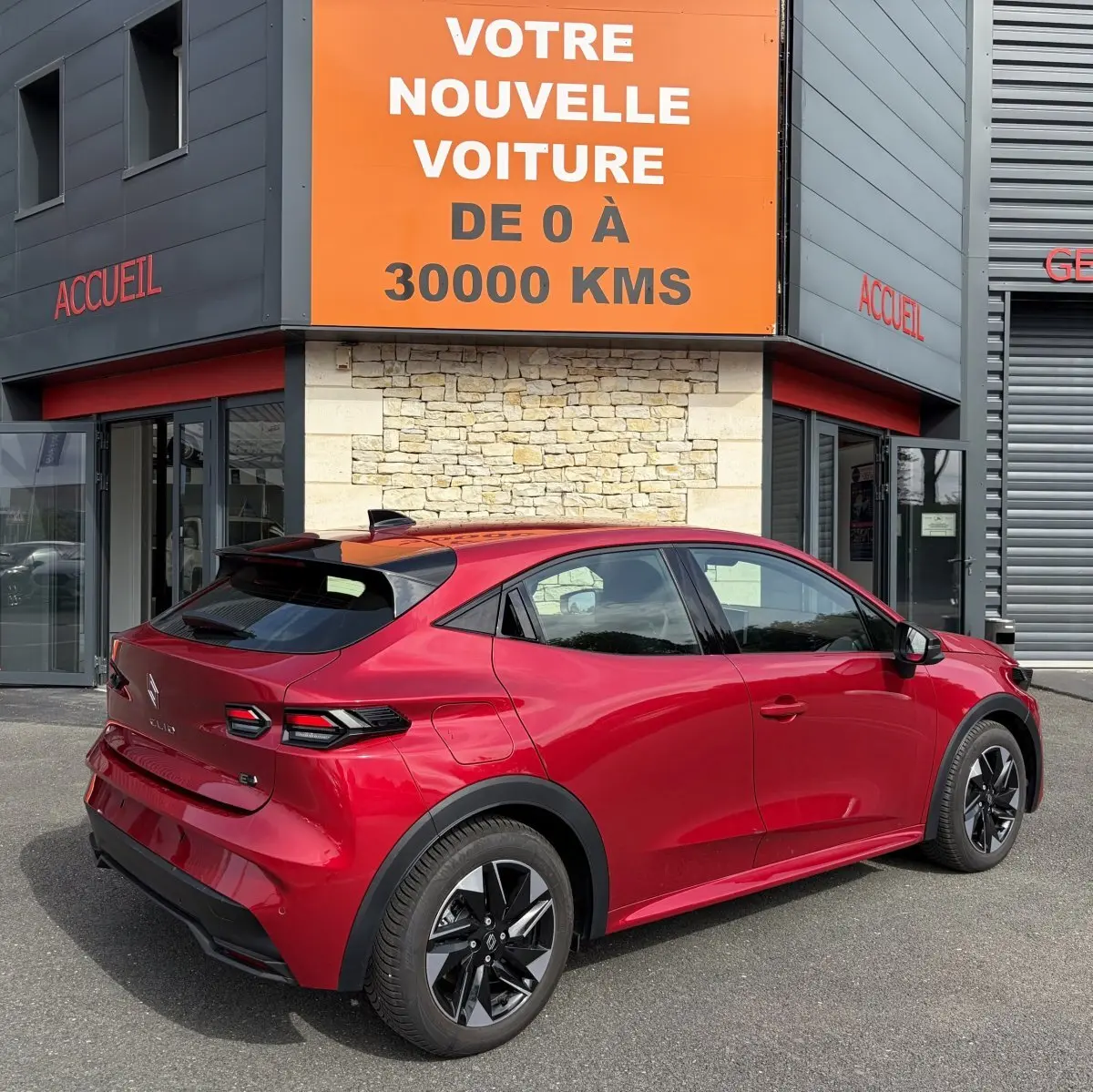 Vue 3/4 arrière droite d'une Renault Clio Evolution rouge 2025 avec toit noir et jantes noires, garée devant un bâtiment commercial.