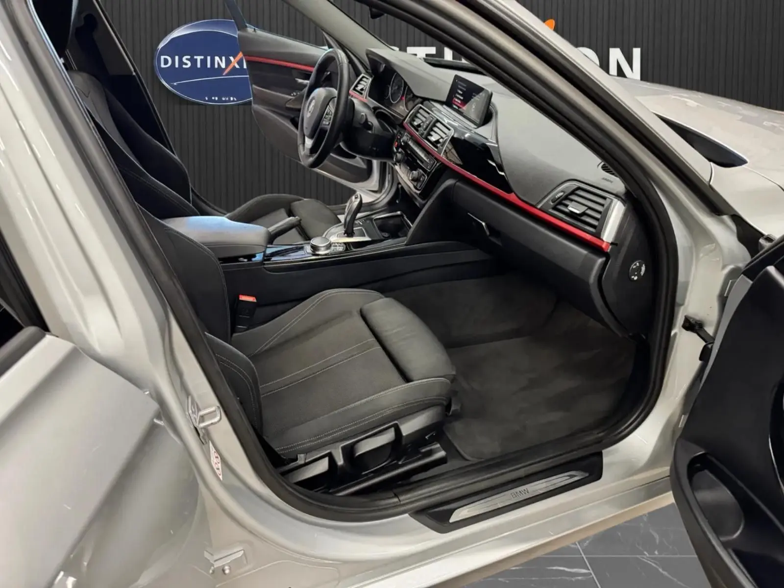 Vue intérieure côté conducteur d'une BMW Série 3 Touring gris aluminium, montrant sièges noirs et tableau de bord avec bande rouge.