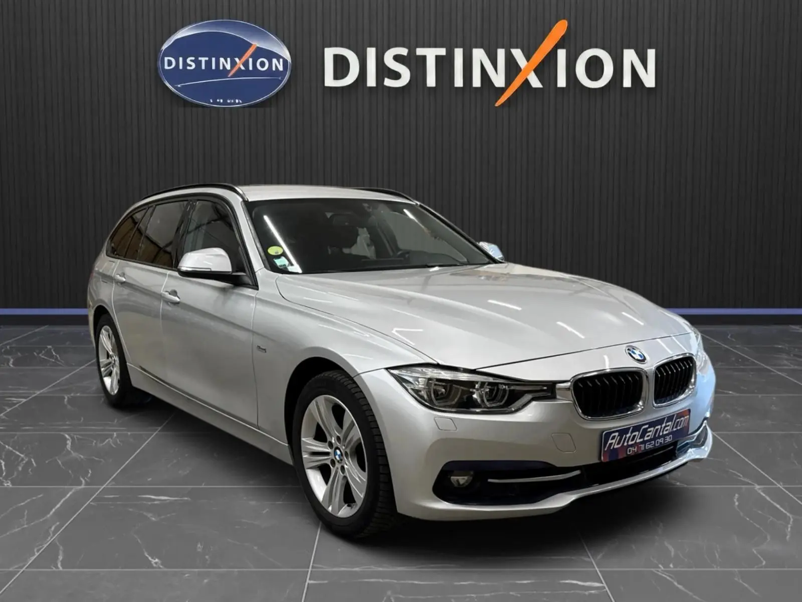 BMW Série 3 Touring F31 gris aluminium vue 3/4 avant droit, stationnée devant un mur noir avec logo Distinxion.
