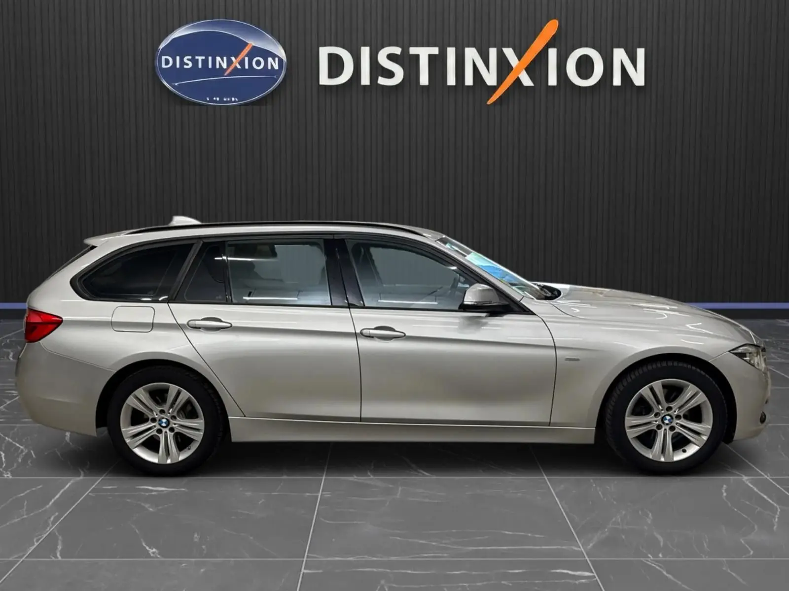 BMW Série 3 Touring F31 gris aluminium vue de profil côté gauche, stationnée devant un mur noir avec logo Distinxion.