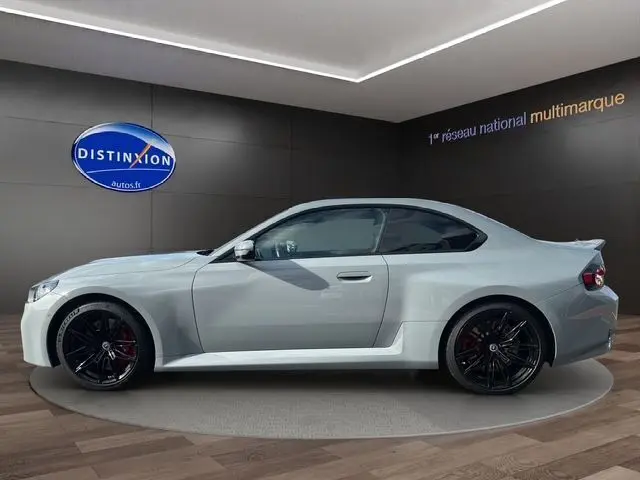 Profil droit d'une BMW M2 G87 gris Brooklyn avec jantes noires et détails sportifs en showroom.