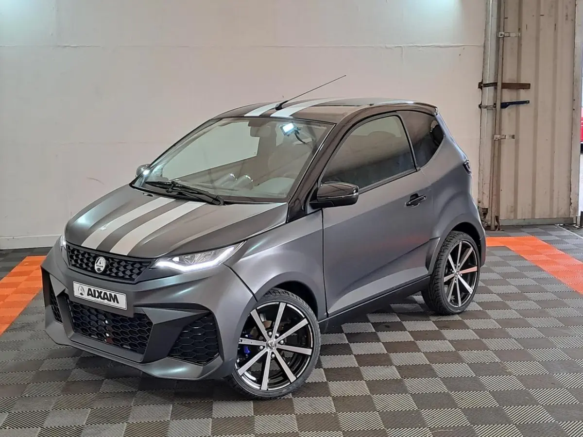 Vue 3/4 avant droite d'une Aixam Ambition gris clair avec bandes blanches, jantes alliage noires diamantées et clignotants à défilement.