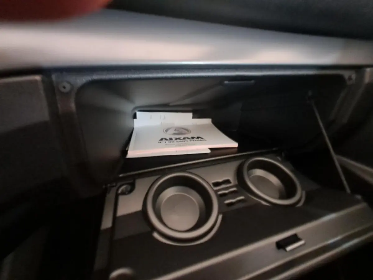 Intérieur de la boîte à gants ouverte de l'AIXAM Ambition 2024 avec porte-gobelets et documentation visible.