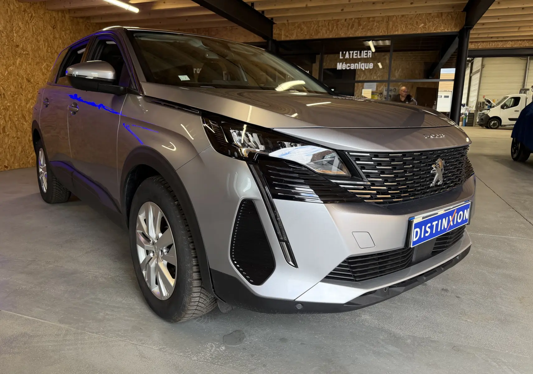 Peugeot 5008 gris vue 3/4 avant droit, avec calandre noire distinctive et phares LED, stationné en intérieur.