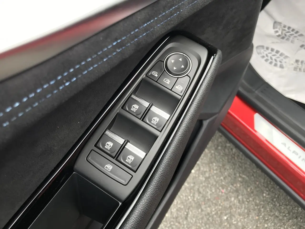 Gros plan sur la commande des vitres côté conducteur avec surpiqûres bleues et seuil de porte rouge Alpine visible.