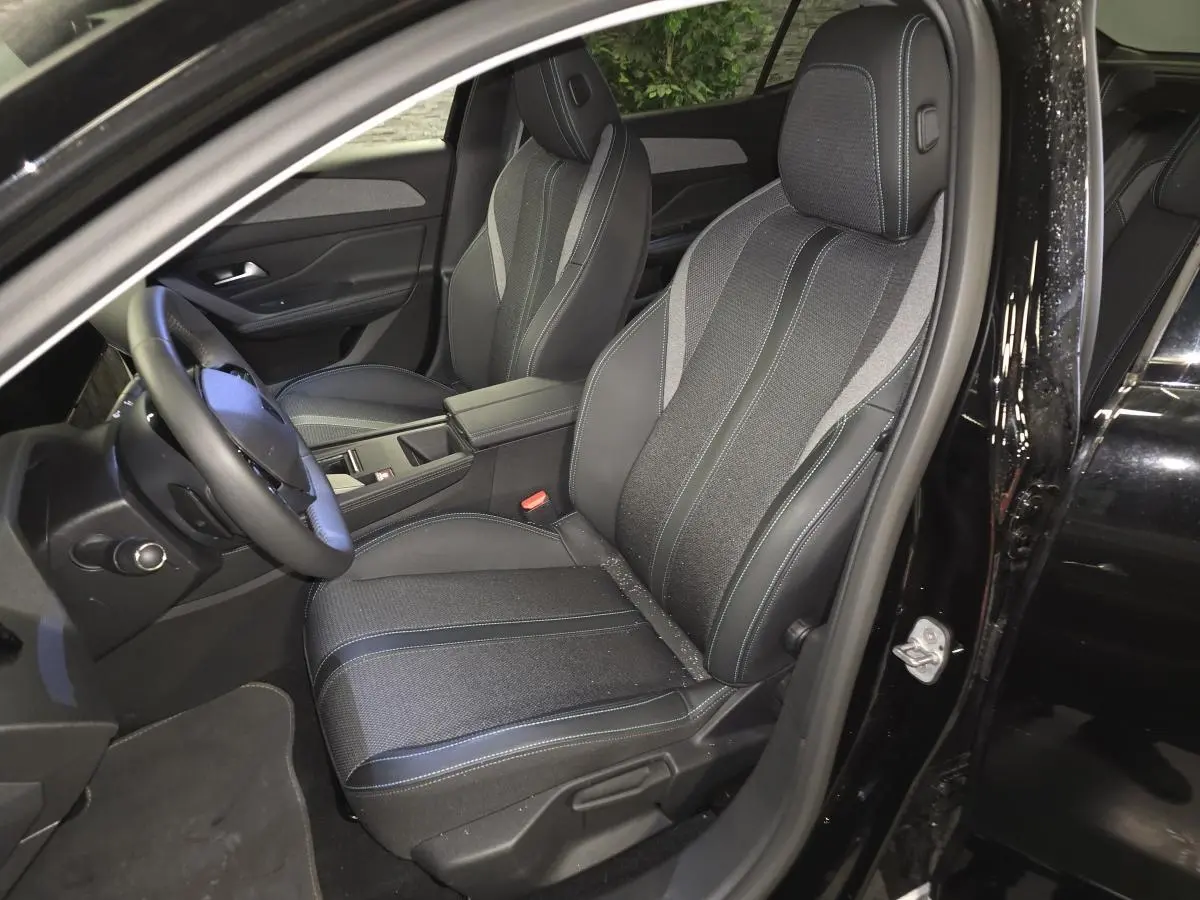 Vue intérieure côté conducteur du Peugeot 408 noir 2024, sièges tri-matières avec surpiqûres bleues et accoudoir central.