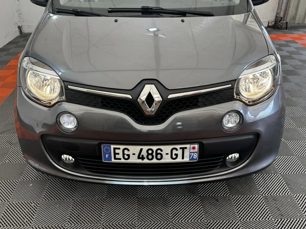 Vue frontale rapprochée d'une Renault Twingo gris foncé avec phares allumés et logo chromé bien visible.