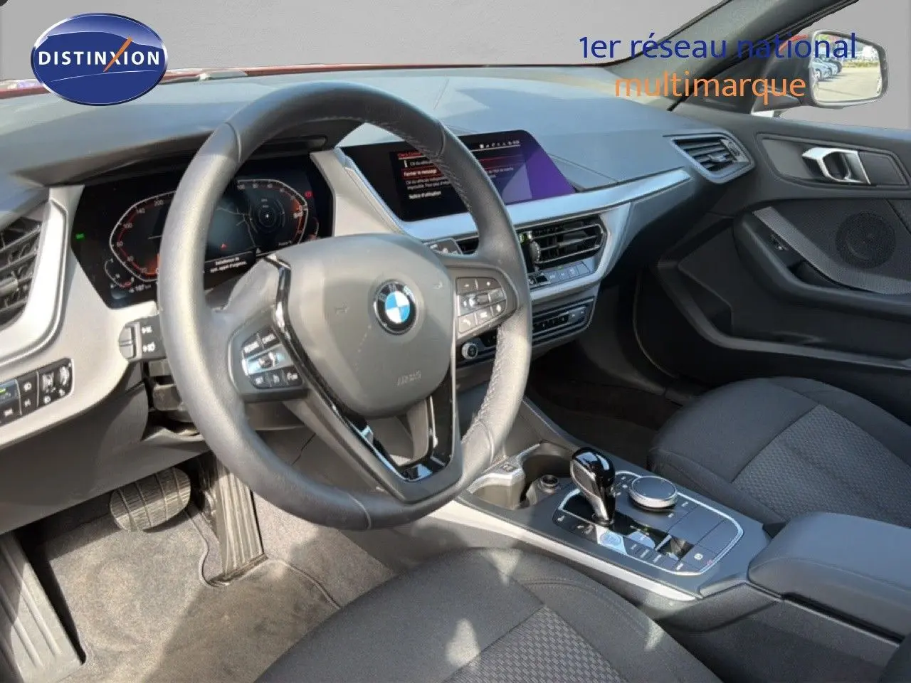 Vue intérieure côté conducteur de la BMW Série 1 116i 2022, volant cuir, console centrale avec boîte auto et écran tactile.
