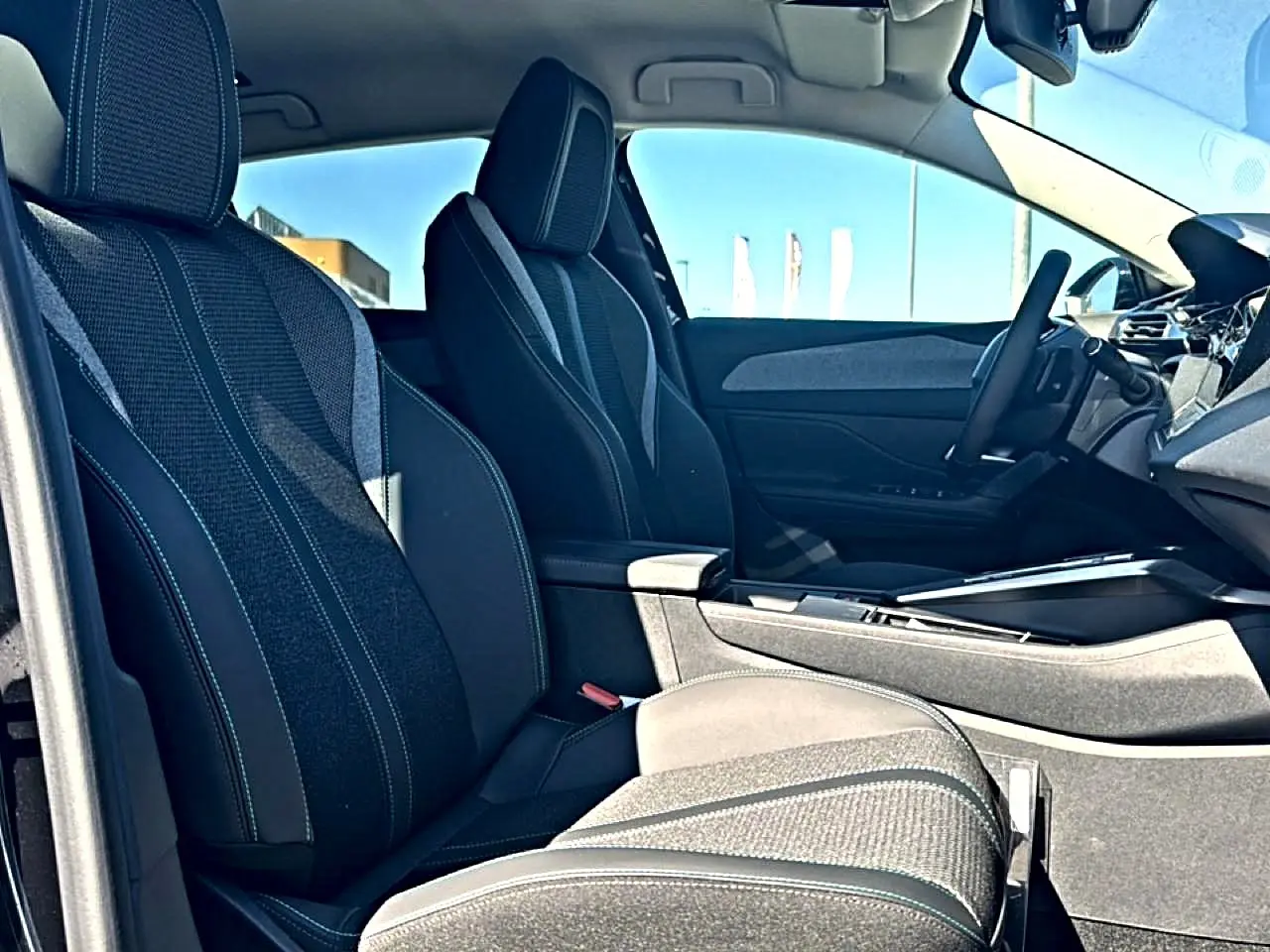 Vue intérieure côté gauche des sièges avant noirs avec accoudoir central et volant cuir de la Peugeot 408 Hybrid 145 allure 2025.