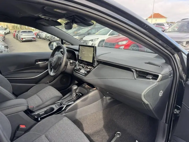 Vue intérieure côté conducteur de la Toyota Corolla 1.8 Hyb noire, avec écran tactile central et sièges sport gris.