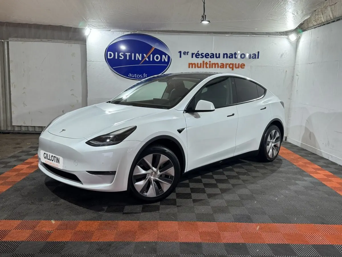 Tesla Model Y blanc en vue 3/4 avant droit, stationné sur sol à damier noir et orange en intérieur showroom.