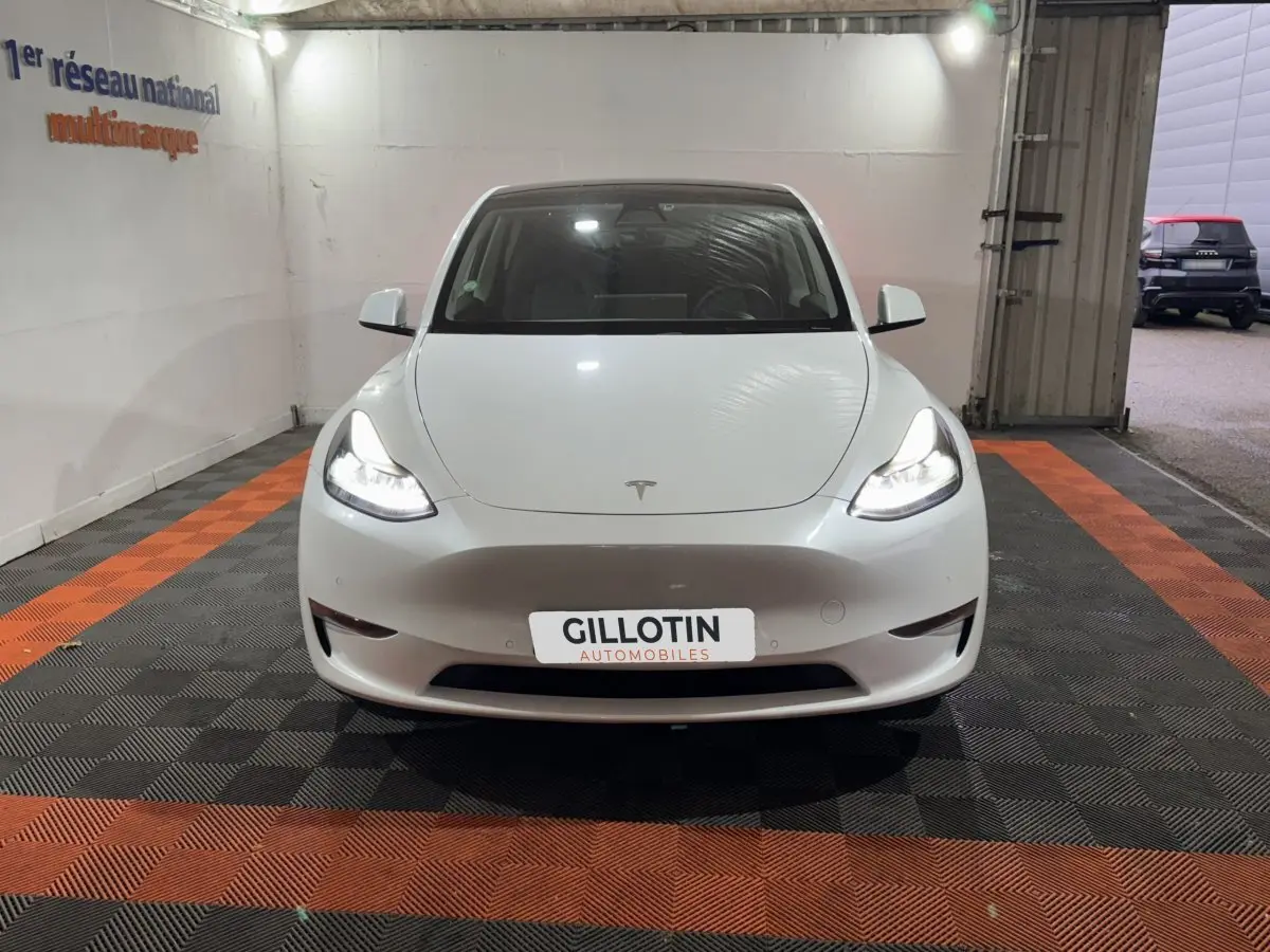 Vue avant d'une Tesla Model Y blanche avec phares allumés dans un garage éclairé.