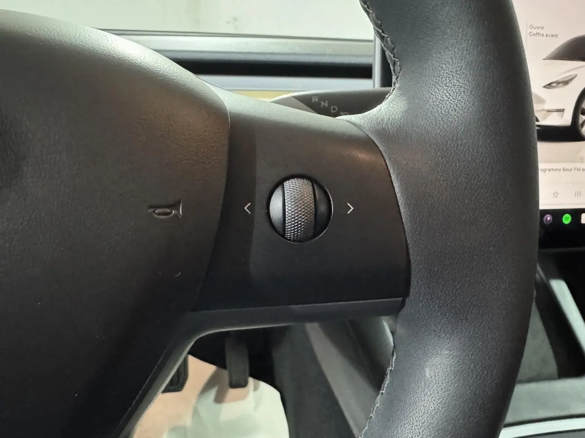 Gros plan sur le volant noir en cuir synthétique de la Tesla Model Y blanc, avec molette de contrôle tactile visible.