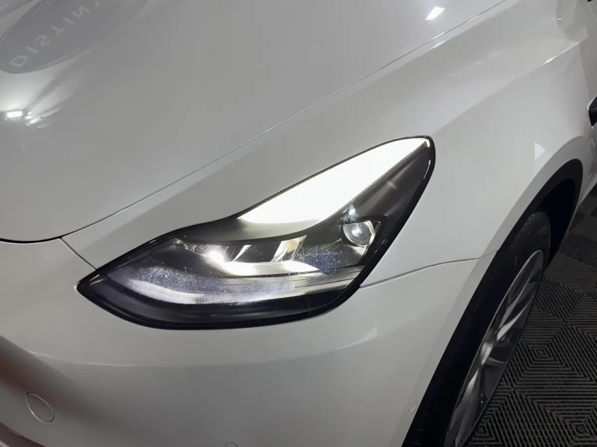 Gros plan sur le phare avant droit allumé d'une Tesla Model Y blanc nacré, avec partie de la roue visible.