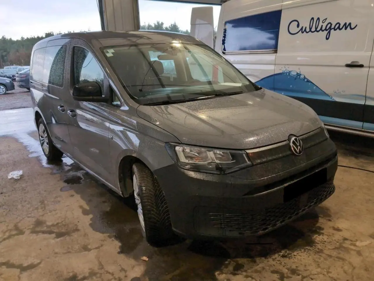 Vue 3/4 avant gauche d'un Volkswagen Caddy Maxi 2024 gris Pure Grey avec portes latérales coulissantes, stationné en intérieur.