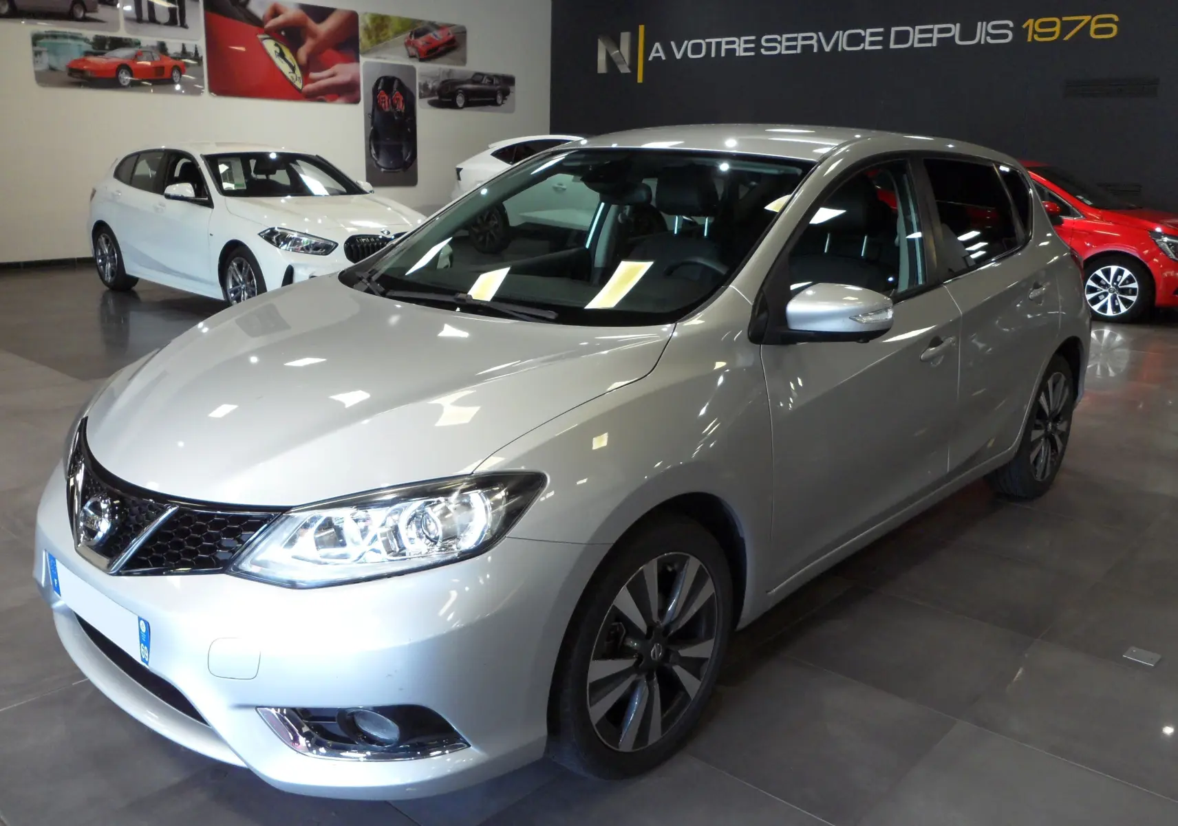 Nissan Pulsar gris glacier vue 3/4 avant droit en intérieur, avec jantes alliage et phares allumés.