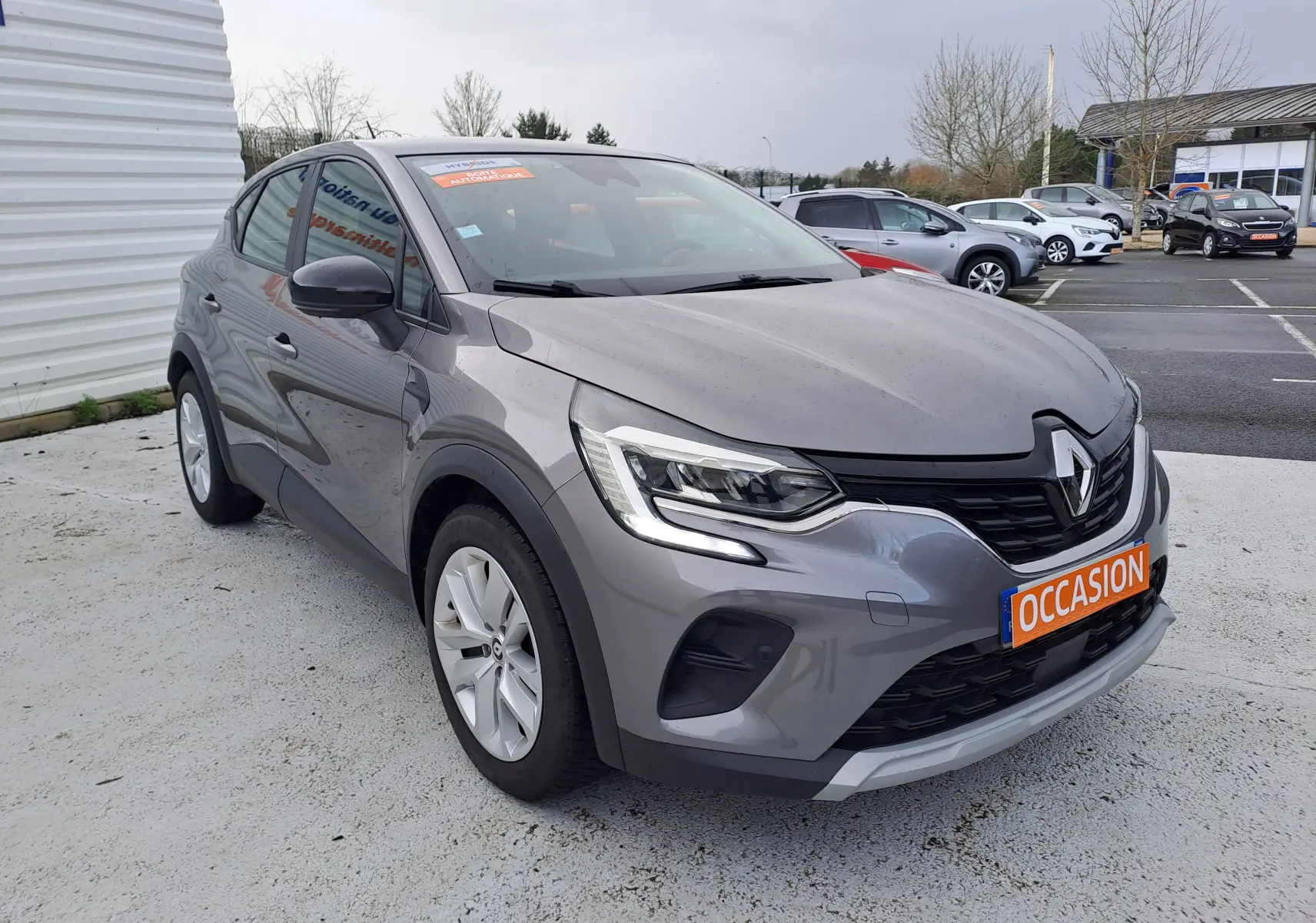 Vue 3/4 avant droite d’un Renault Captur gris cassiopée métal avec plaque orange « OCCASION » sur un parking extérieur.