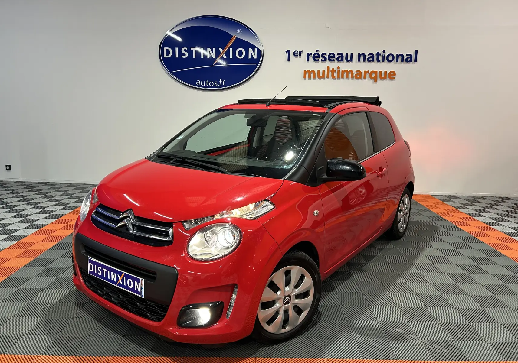 Citroën C1 rouge vue 3/4 avant droit, toit ouvrant Airscape ouvert, dans un showroom avec sol gris et orange.