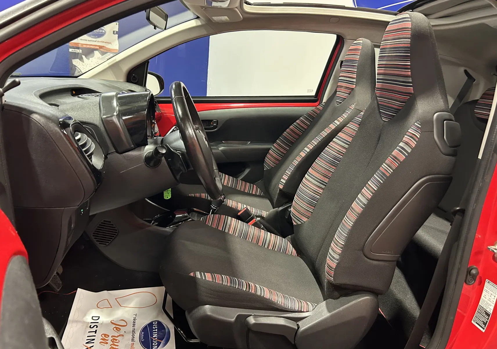 Vue intérieure côté conducteur d'une Citroën C1 rouge 2019, sièges tissu rayé noir et rouge, tableau de bord compact.