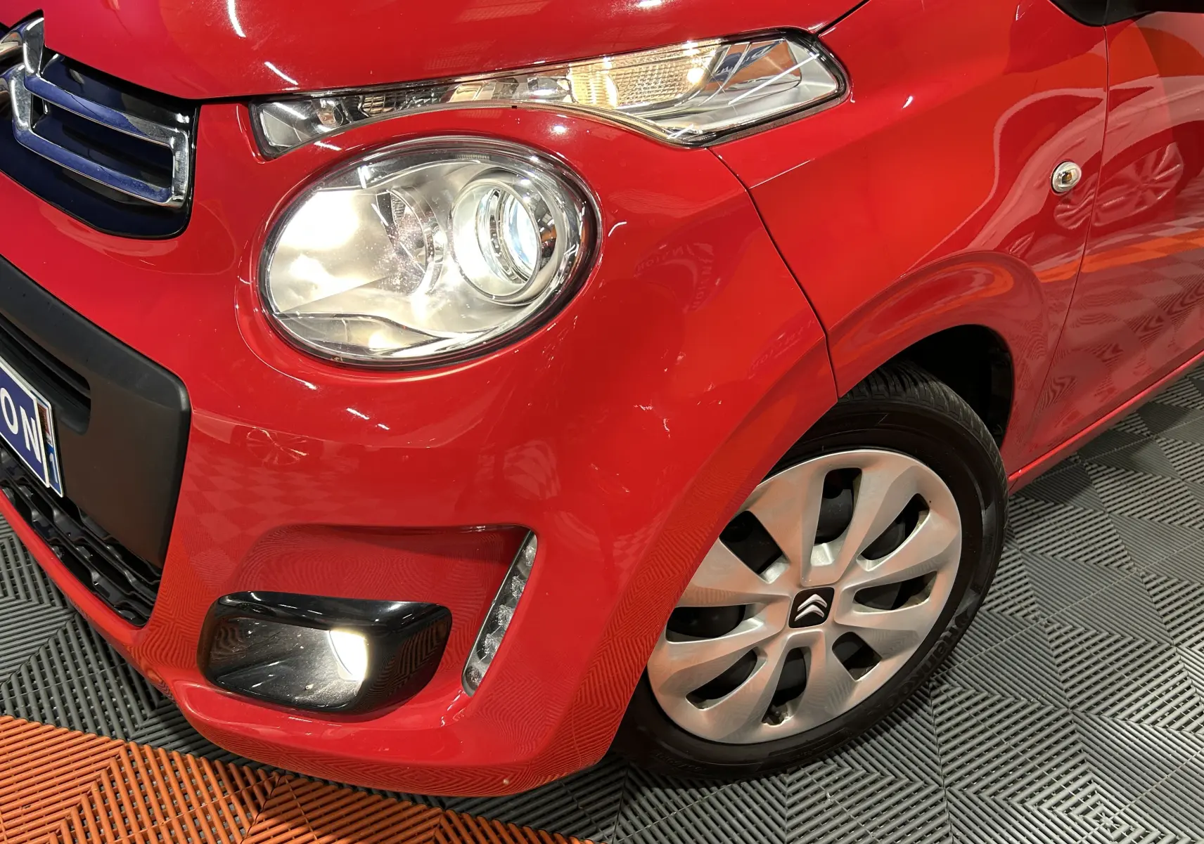 Gros plan sur l’avant droit d’une Citroën C1 rouge 2019 avec phare allumé et jante alu distinctive.