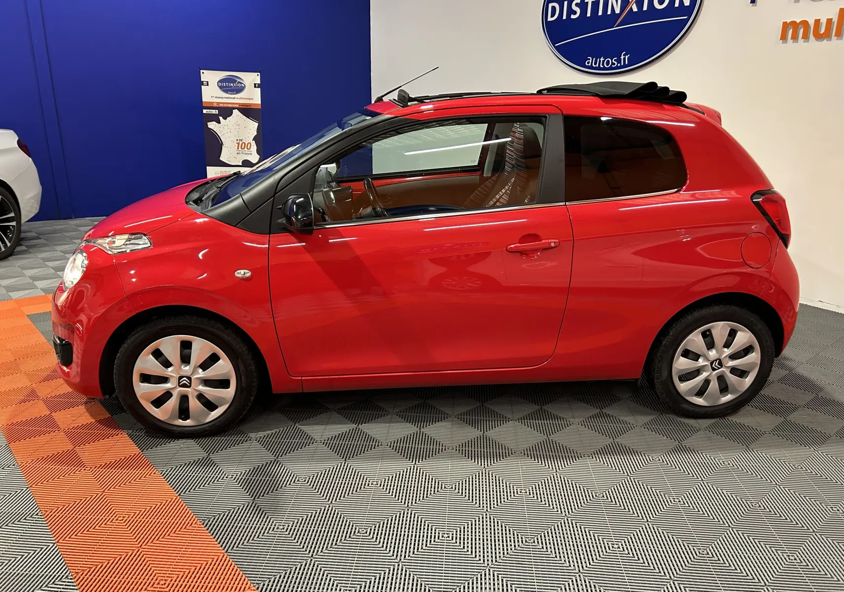 Vue de profil côté gauche d'une Citroën C1 Airscape rouge avec toit ouvrant en toile replié, dans un showroom.