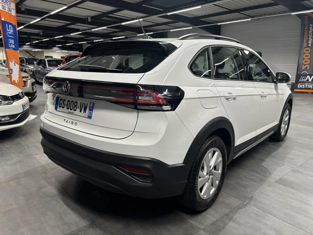 Vue 3/4 arrière droite d’un Volkswagen Taigo blanc avec feux arrière noirs et jantes alliage visibles en intérieur showroom.