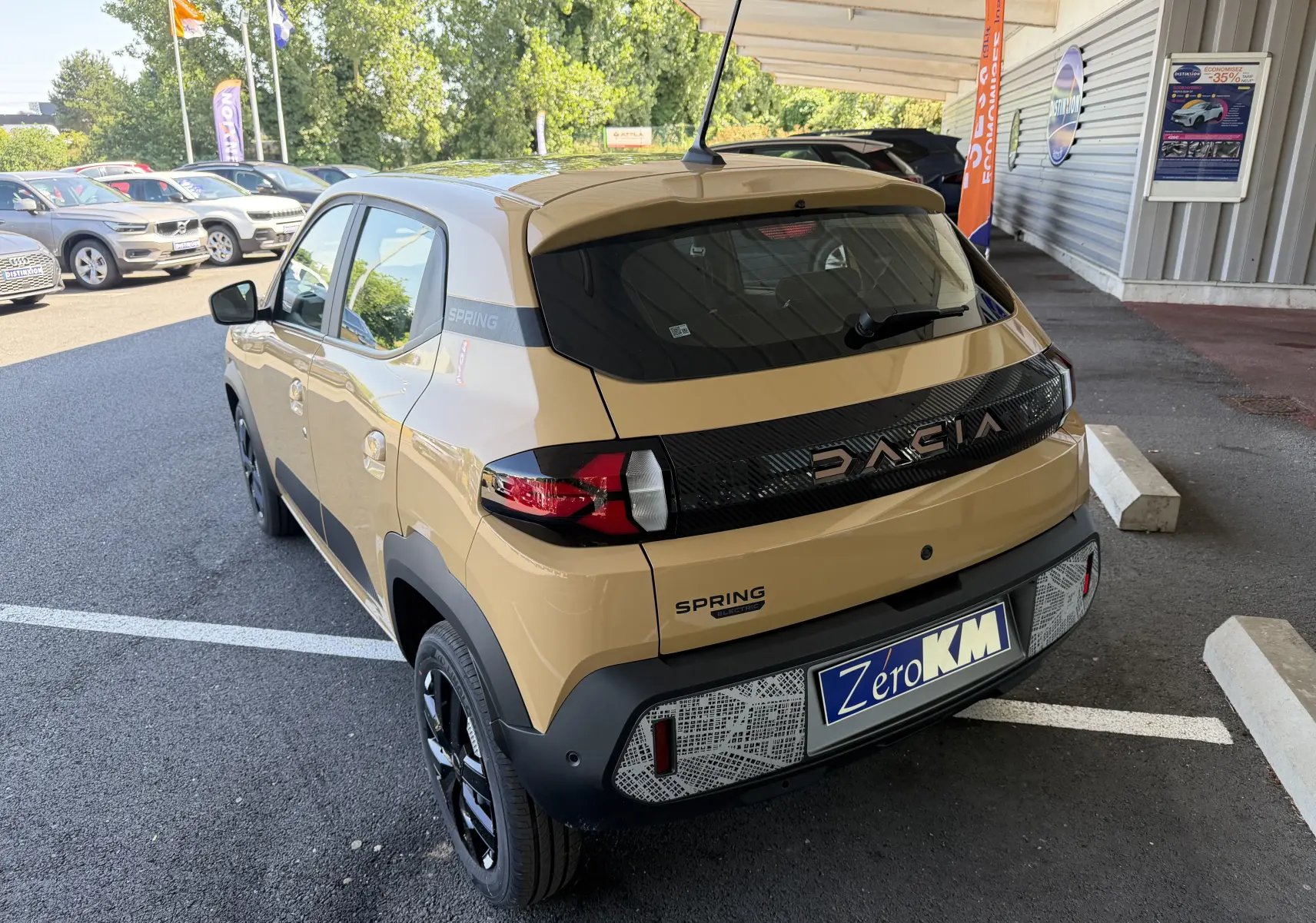 Vue 3/4 arrière droite d'une Dacia Spring 65 Extreme beige Safari avec logo DACIA noir brillant et plaque Zéro KM.