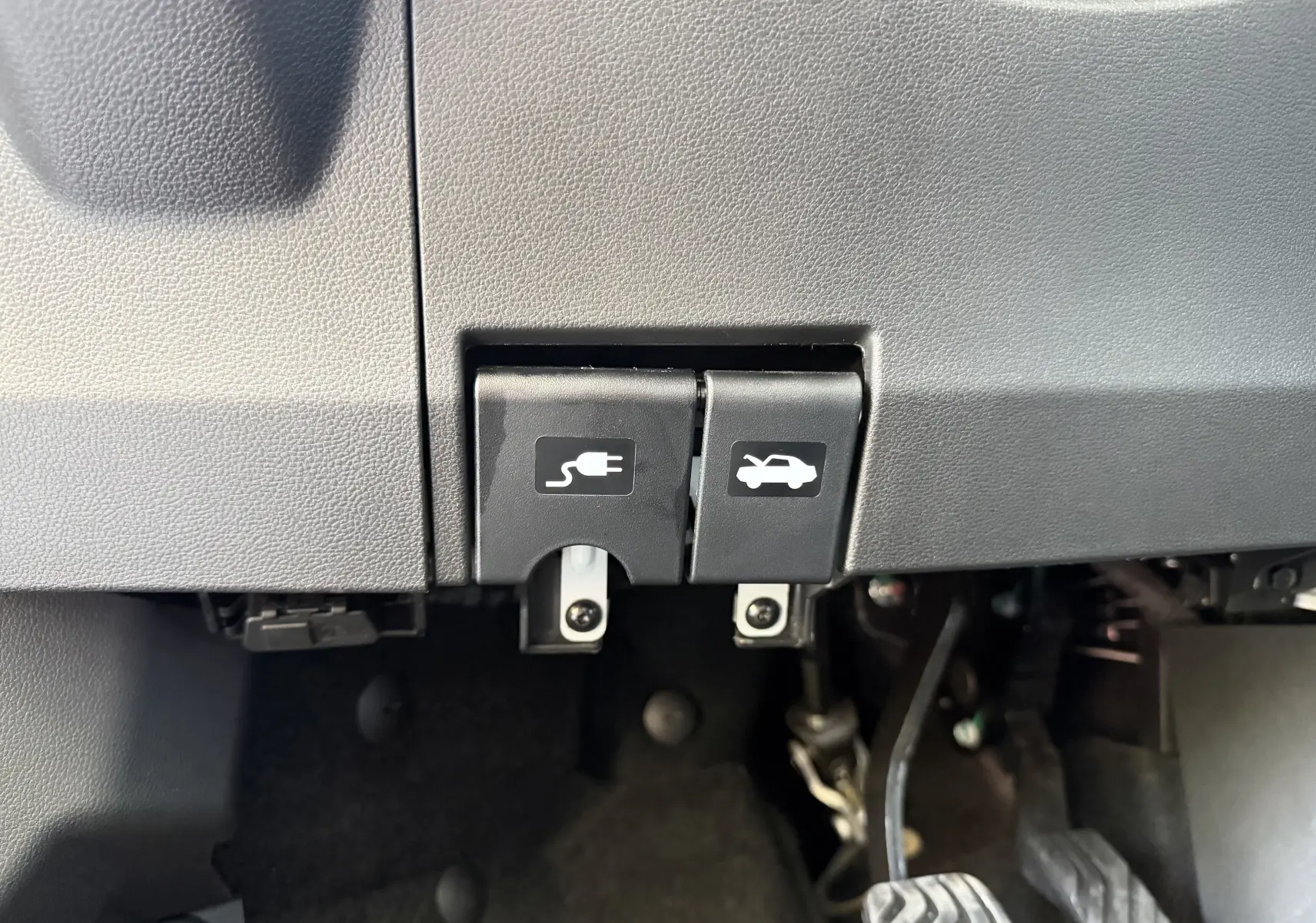 Gros plan sur les commandes d’ouverture de capot et de trappe de recharge électrique sous le tableau de bord d’une Dacia Spring beige Safari.