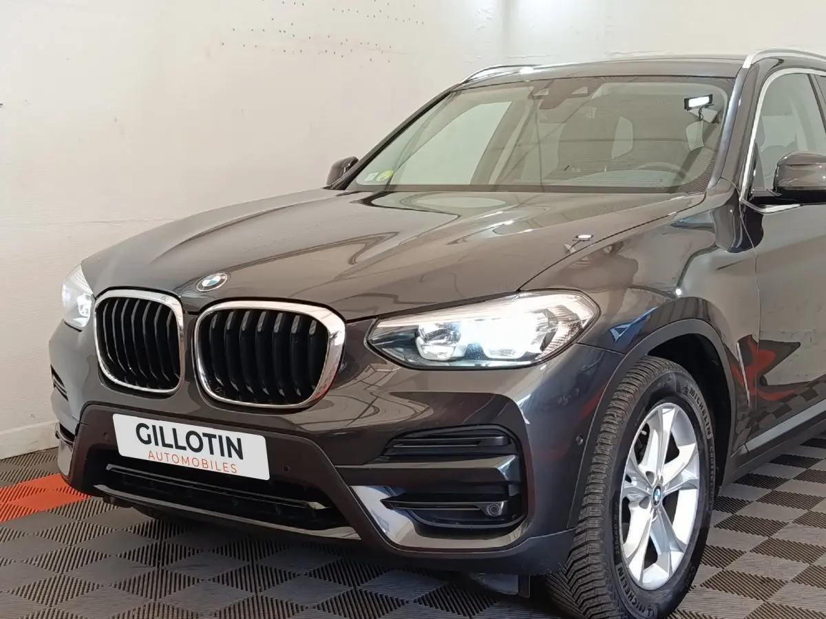 Vue 3/4 avant droite d’un BMW X3 gris clair avec calandre double haricot et phares allumés en intérieur showroom.