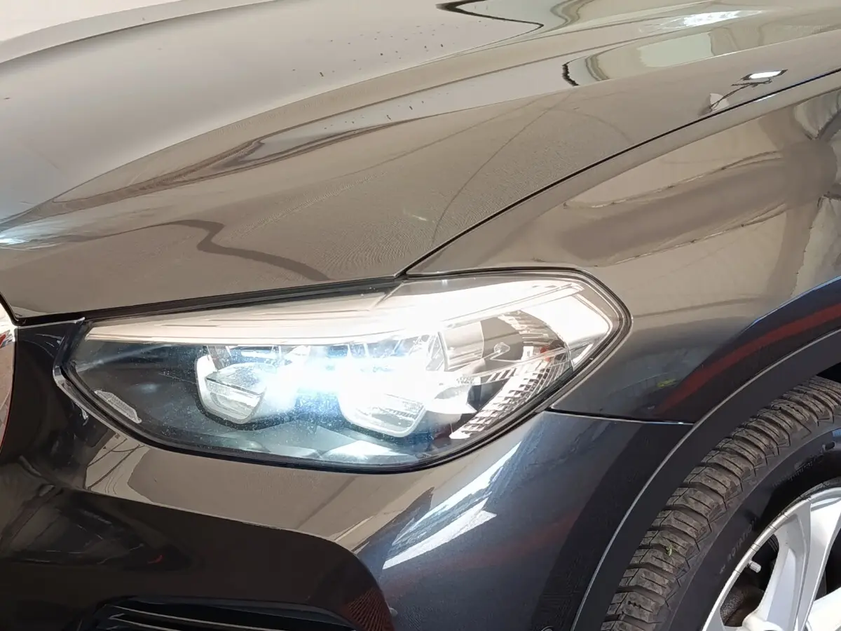 Gros plan sur le phare avant droit allumé d’un BMW X3 gris clair avec jante alu visible.