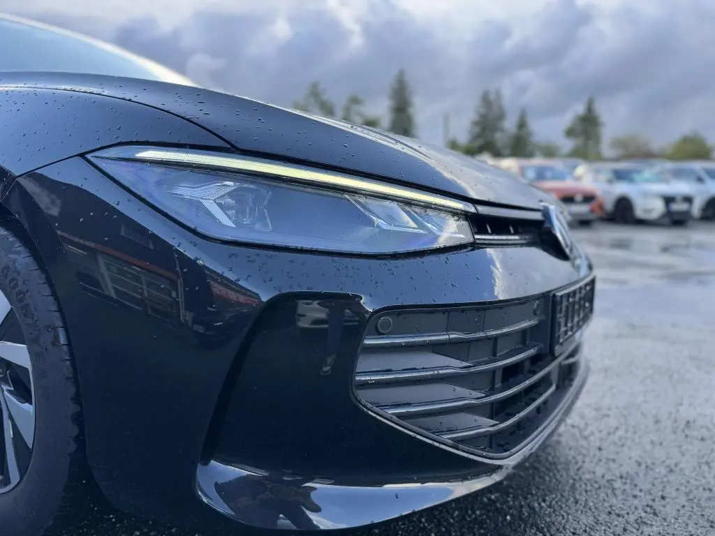 Gros plan sur le phare avant gauche d'une Volkswagen Passat SW noire 2024 avec gouttes de pluie et calandre visible.